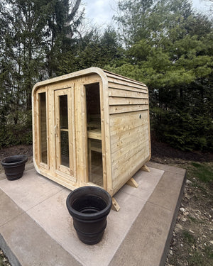 Cedar cabin sauna with a three-panel glass front on a tiered concrete pad, backed by tall evergreens