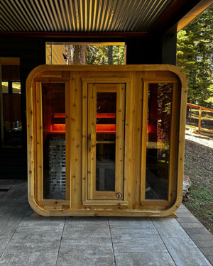 Cedar cabin sauna with three glass front panels glowing red inside, under a covered patio beside a pine forest