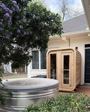 Cedar cabin sauna on a brick patio beside a round stock-tank plunge under a purple-flowering tree