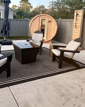 Cedar barrel sauna and cedar outdoor shower at the edge of a fenced patio with lounge chairs, a rug and a fire table