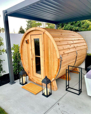 Cedar barrel sauna under a modern louvered pergola with lanterns, a slatted mat and a towel rack