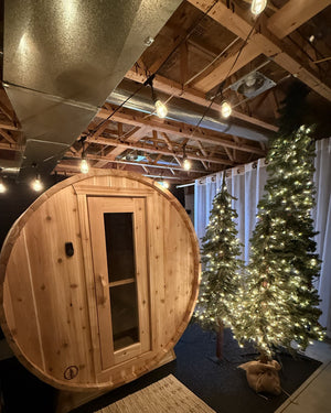 Cedar barrel sauna in a basement under exposed joists and string lights, beside two lit artificial pine trees