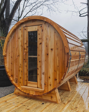 Rain-wet cedar barrel sauna on a new wooden deck in a fenced yard, with bare trees and water beyond