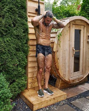 Man rinsing off under a cedar outdoor shower beside a cedar barrel sauna in a hedged garden