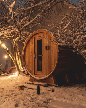 Cedar barrel sauna lit warm at night beneath snow-covered birch branches in a snowy backyard