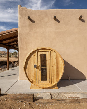 Cedar barrel sauna on a concrete pad against an adobe-style stucco wall, with cacti and desert hills under a blue sky