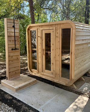 Cedar cabin sauna with three glass front panels next to a cedar outdoor shower in a wooded yard