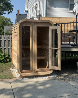 Cedar cabin sauna with its glass door open on a gravel pad beside a backyard deck