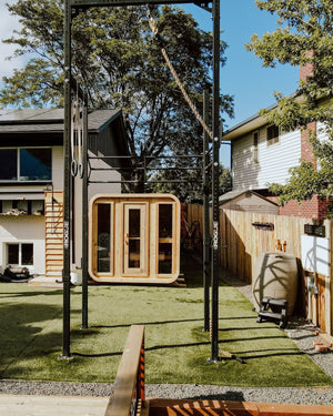 Cedar cabin sauna with three glass front panels in a turf backyard gym framed by a pull-up rig