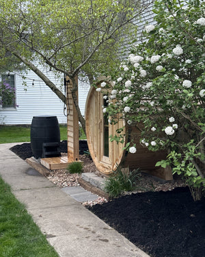 Barrel sauna and cedar outdoor shower tucked behind a white flowering shrub in a mulched garden bed