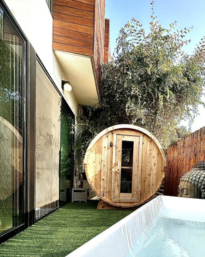 Cedar barrel sauna on artificial turf between a modern house and a bamboo fence, with a white tub in front