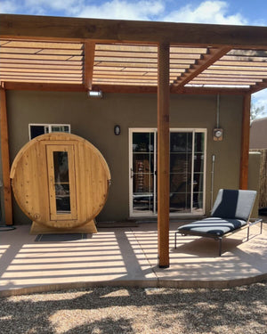 Cedar barrel sauna under a wood pergola on a stucco home's patio, beside a lounge chair and sliding door