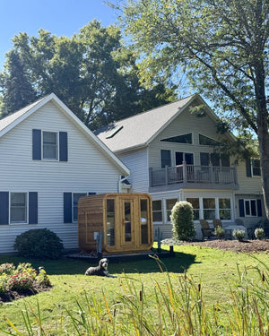 Cedar cabin sauna on a sunny lawn behind a large white house, with a dog resting on the grass