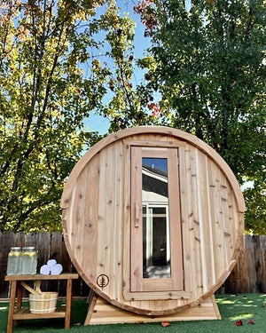 Cedar barrel sauna on artificial turf with a side table of drinks, towels and a cedar bucket under autumn trees
