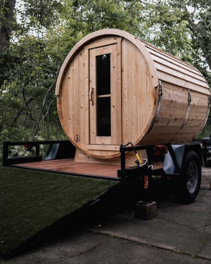 Cedar barrel sauna mounted on a flatbed trailer with a turf-covered ramp, parked at the edge of a forest