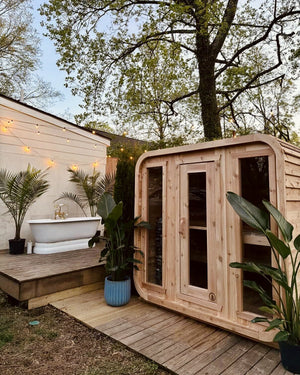 Cedar cabin sauna on a wooden deck at dusk, with string lights, potted palms and a white clawfoot tub