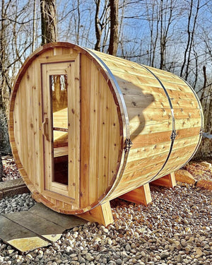 Cedar barrel sauna on a river-rock pad in bare winter woods, lit by warm low sun