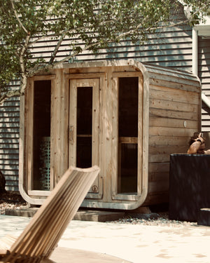 Cedar cabin sauna with three glass front panels, a cedar outdoor shower and a man in a black cold plunge on a patio