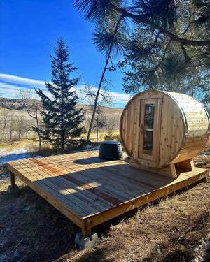 Cedar barrel sauna on a new wood deck among pines, overlooking open hills with patches of snow