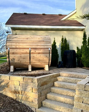 Long cedar barrel sauna seen side-on on a paver pad behind a house, above stone steps and a retaining wall