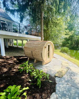 Cedar barrel sauna on a flagstone path beside a calm pond, beneath a raised house deck