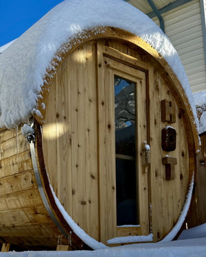 Cedar barrel sauna heaped with fresh snow under a clear blue sky, seen from ground level