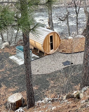 Long cedar barrel sauna and a stock tank plunge beside a gravel path and a rushing river, seen through pines