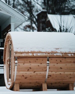 Snow-covered cedar barrel sauna in a backyard during a snowfall, with a deck and string lights blurred behind