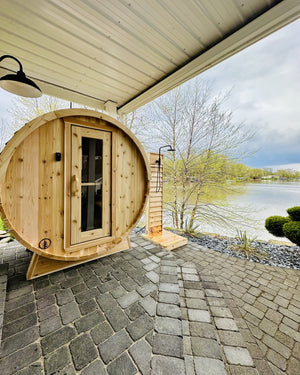Cedar barrel sauna and cedar outdoor shower on a covered paver patio beside a lake with budding trees