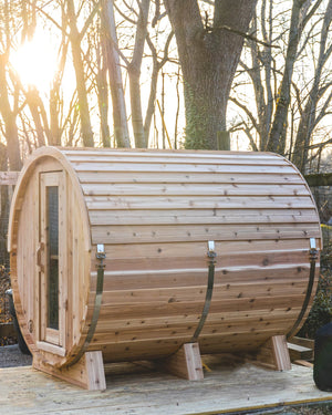 Cedar barrel sauna on a wooden platform at the edge of the woods, sun glowing through bare trees