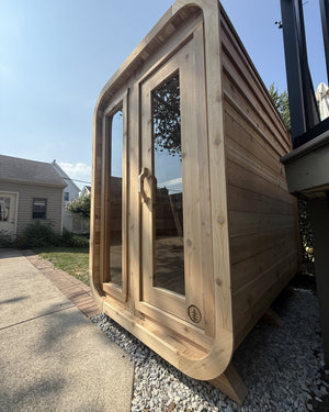 Low-angle view of a rounded cedar cabin sauna with two tall glass panels on a gravel pad under a blue sky