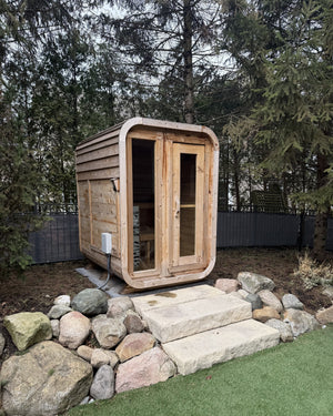 Compact cedar cabin sauna with a glass front among evergreens, reached by stone slab steps from the lawn