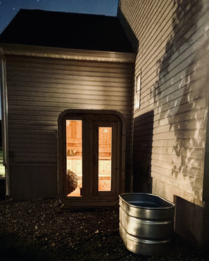 Cabin sauna glowing warm at night against a house wall under the stars, with a metal stock-tank plunge beside it