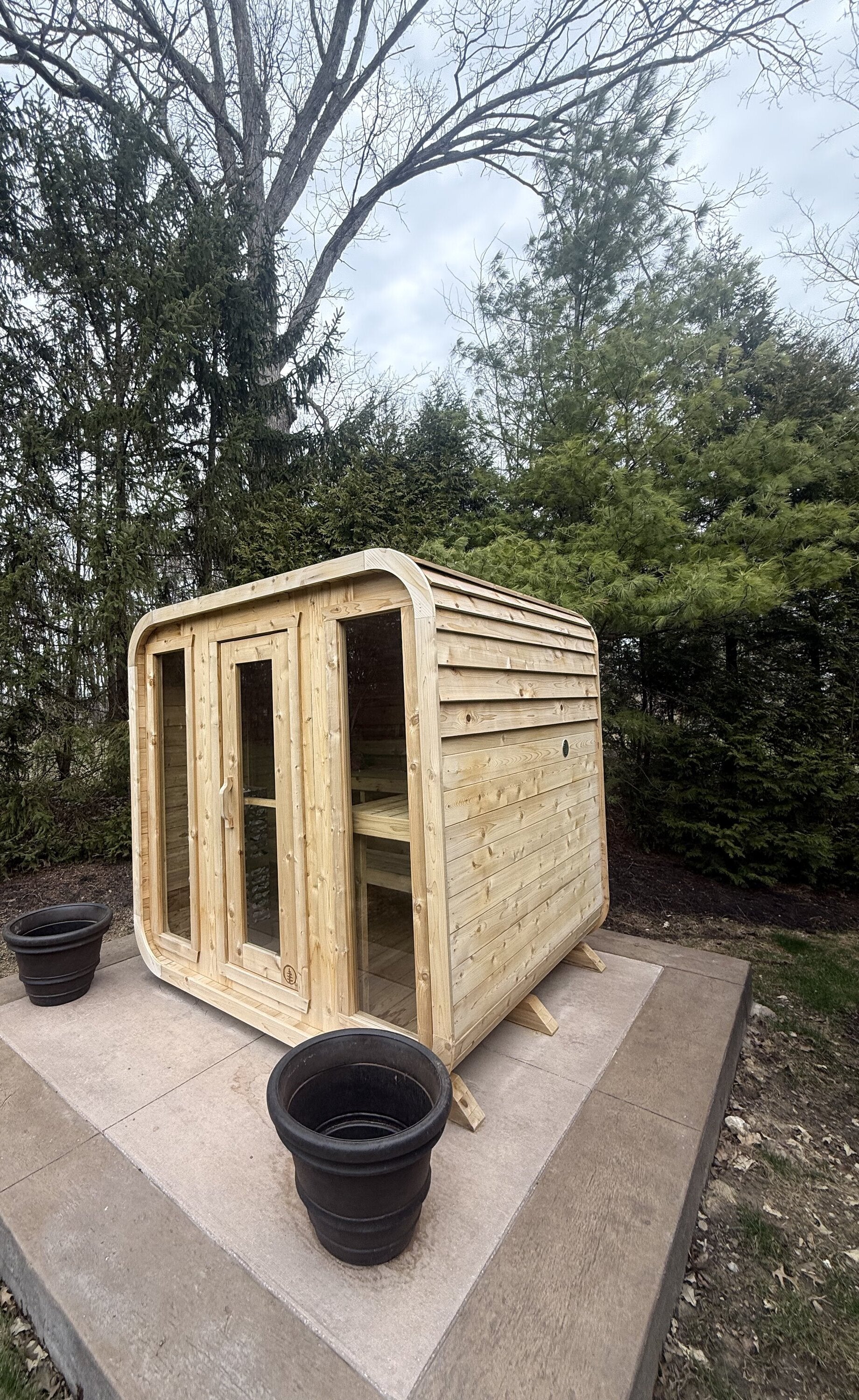 Cedar cabin sauna with a three-panel glass front on a tiered concrete pad, backed by tall evergreens