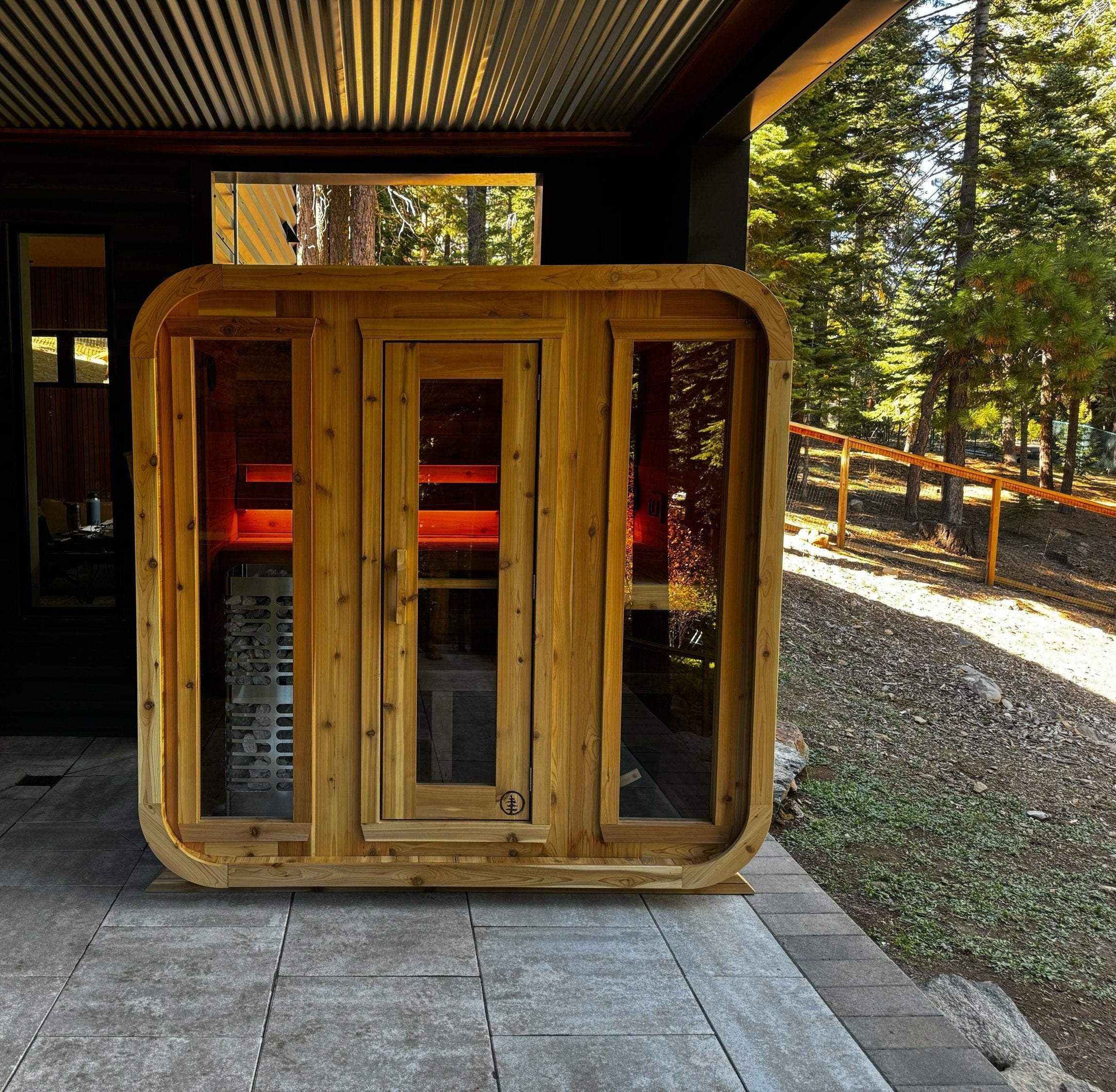 Cedar cabin sauna with three glass front panels glowing red inside, under a covered patio beside a pine forest