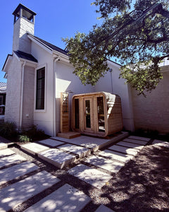 Cedar cabin sauna and outdoor shower on a concrete pad beside a white farmhouse, reached by stepping-stone pavers
