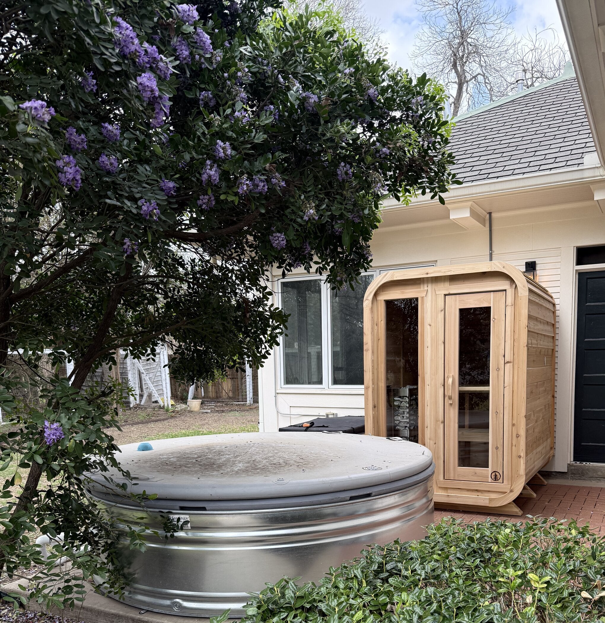 Cedar cabin sauna on a brick patio beside a round stock-tank plunge under a purple-flowering tree