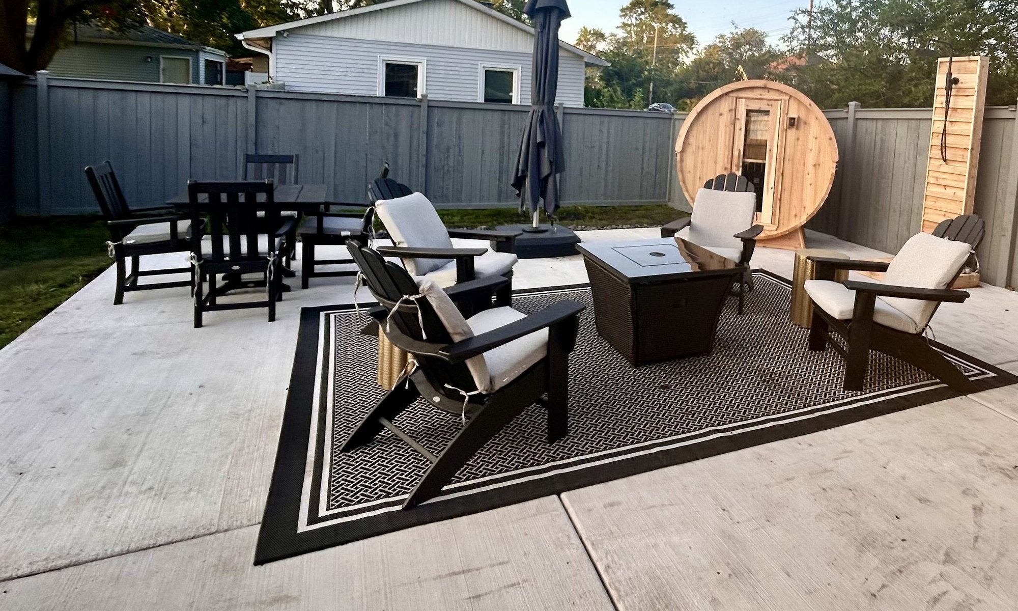 Cedar barrel sauna and cedar outdoor shower at the edge of a fenced patio with lounge chairs, a rug and a fire table