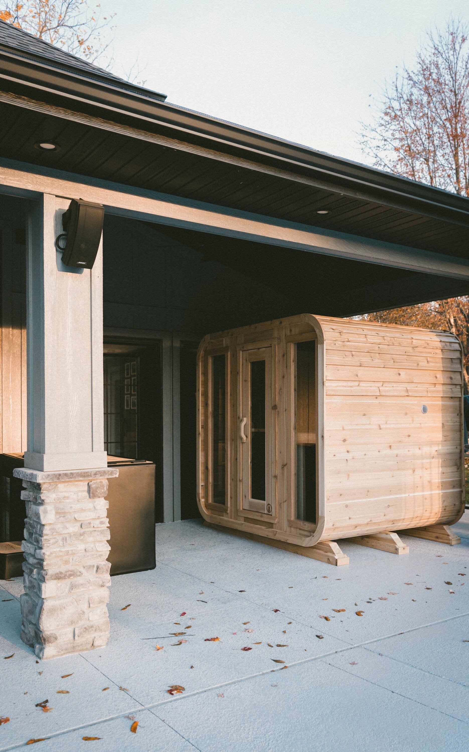 Cedar cabin sauna with three glass front panels under a covered patio in warm autumn light