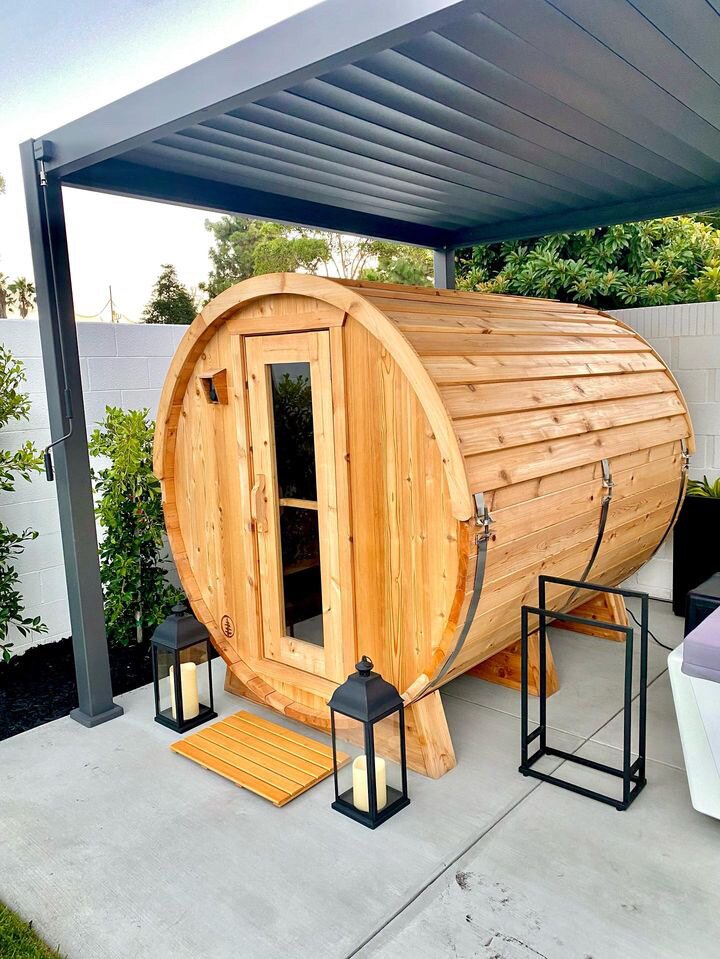 Cedar barrel sauna under a modern louvered pergola with lanterns, a slatted mat and a towel rack