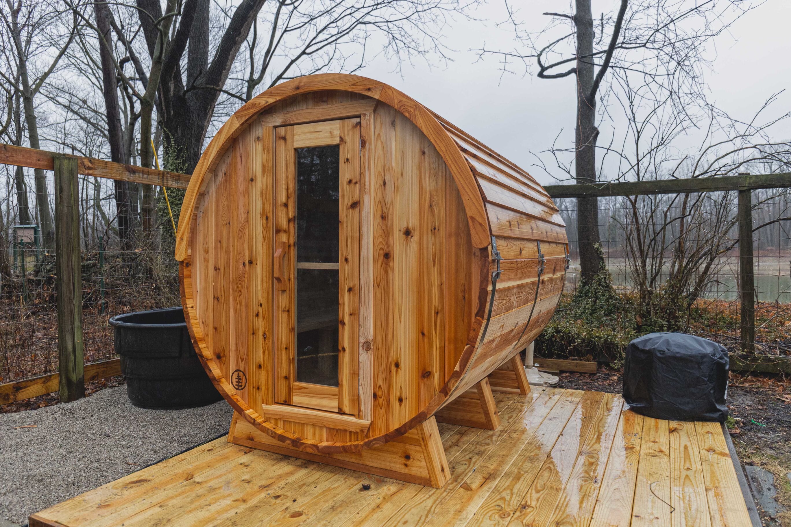 Rain-wet cedar barrel sauna on a new wooden deck in a fenced yard, with bare trees and water beyond