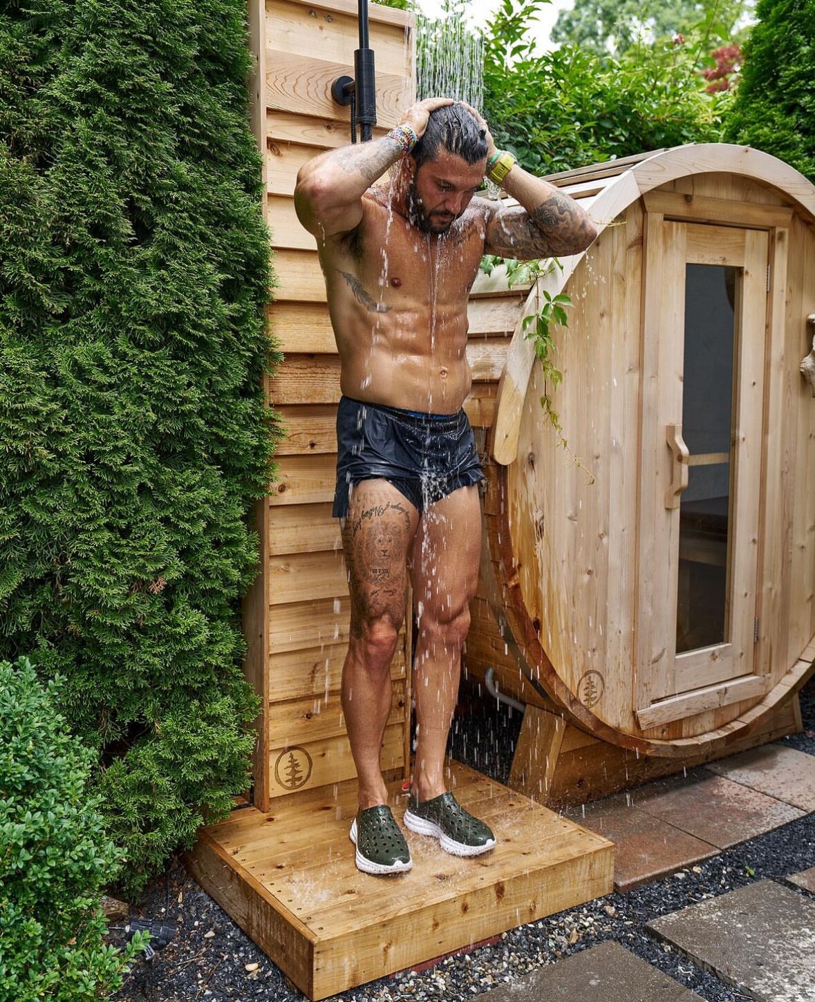 Man rinsing off under a cedar outdoor shower beside a cedar barrel sauna in a hedged garden