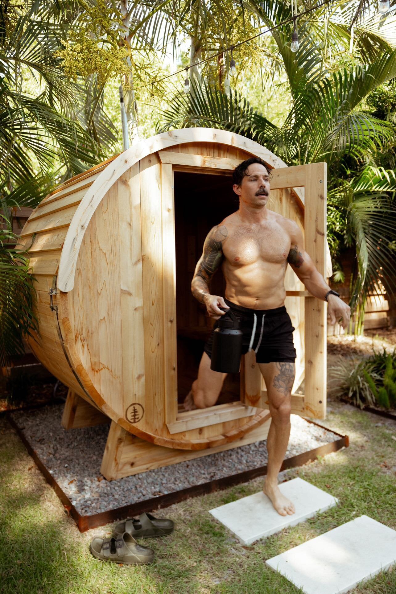 Man with a water jug stepping out of a cedar barrel sauna onto stepping stones in a palm garden