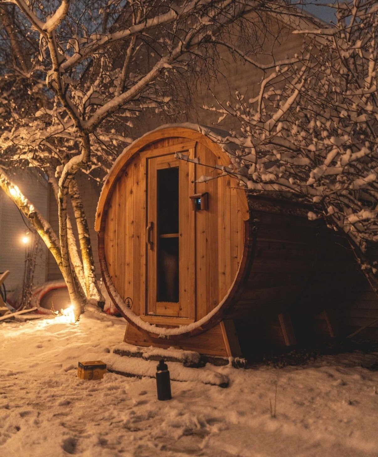 Cedar barrel sauna lit warm at night beneath snow-covered birch branches in a snowy backyard