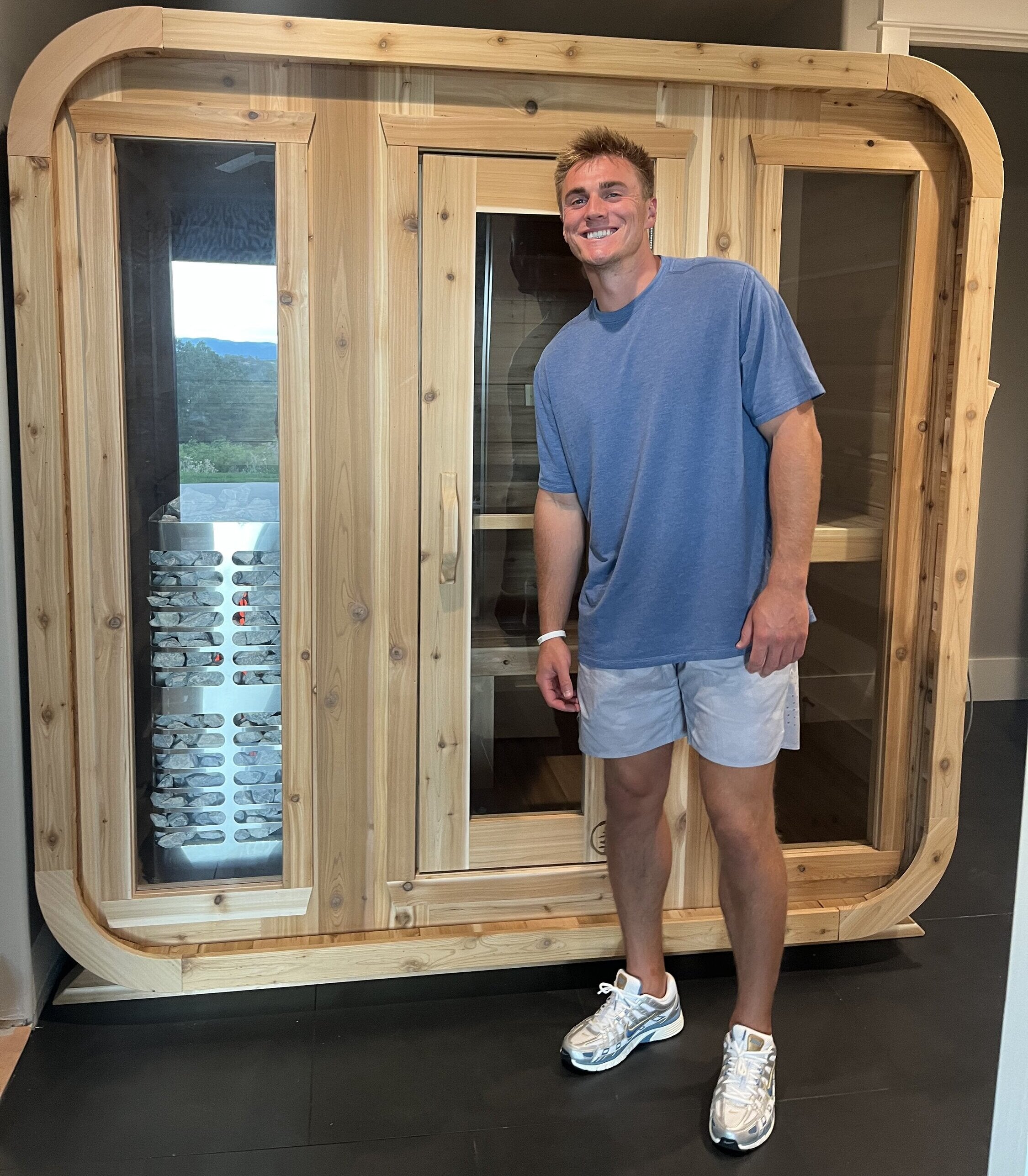 Smiling man standing in front of an indoor cedar cabin sauna with three glass panels