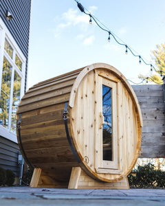 Cedar barrel sauna on a stone patio beside a gray house, under string lights, with a wood screen and pines behind