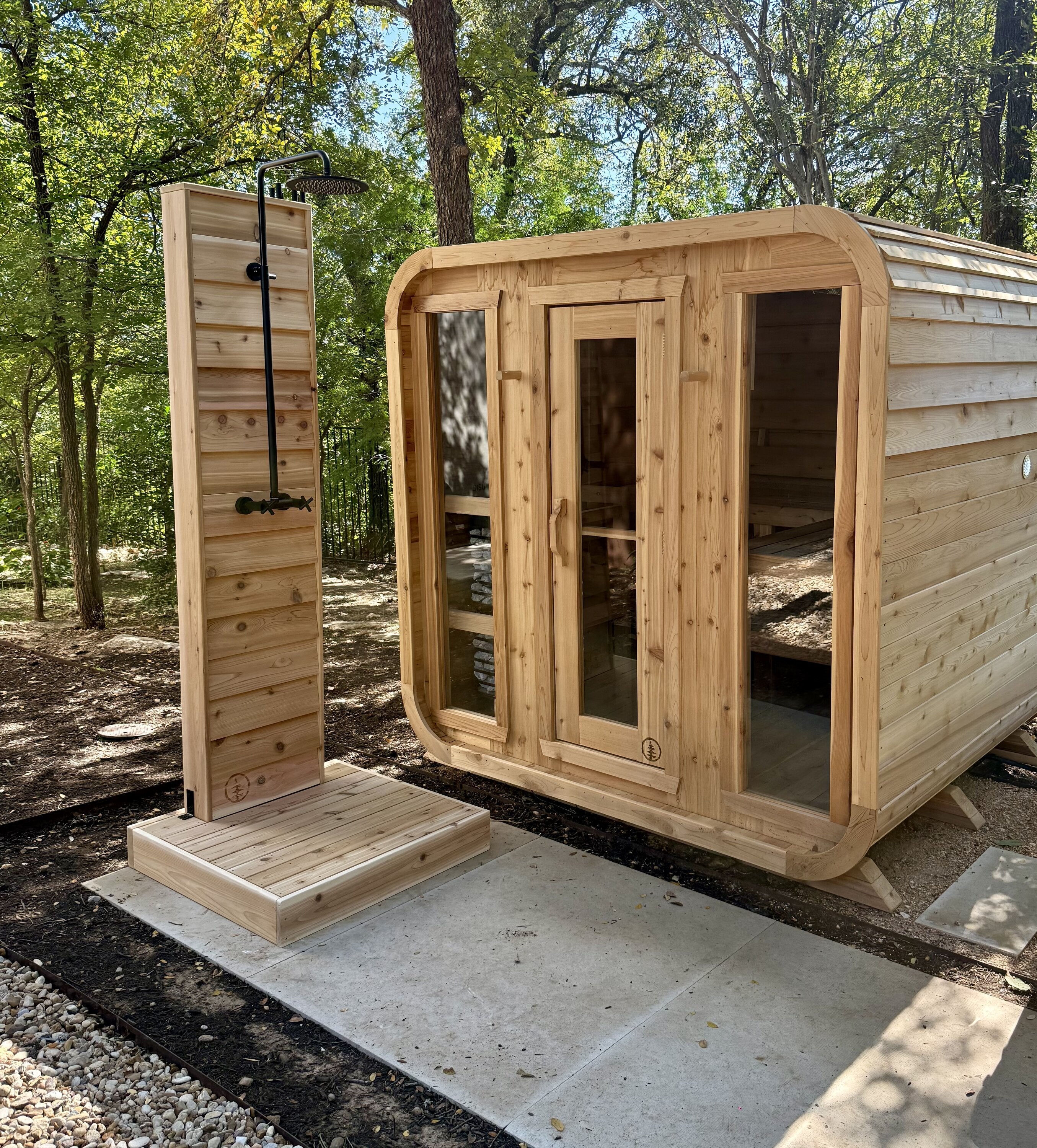Cedar cabin sauna with three glass front panels next to a cedar outdoor shower in a wooded yard