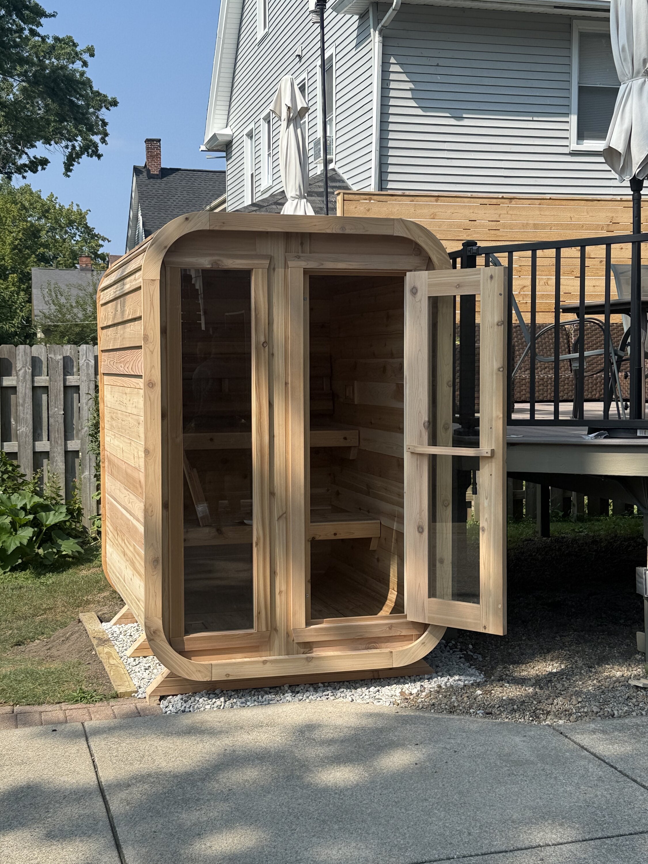 Cedar cabin sauna with its glass door open on a gravel pad beside a backyard deck