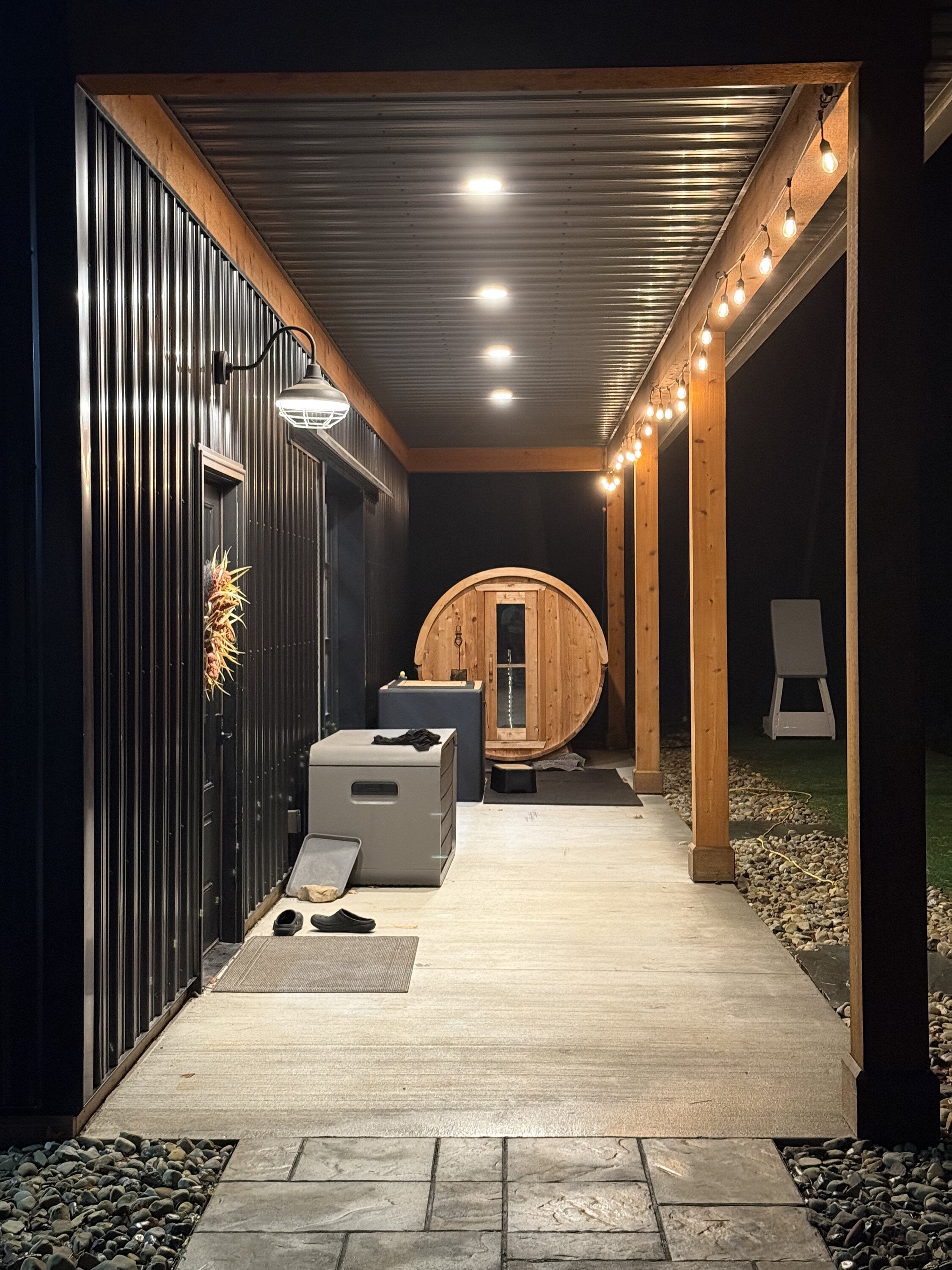 Cedar barrel sauna glowing at the end of a covered walkway lit by string lights and ceiling lights at night