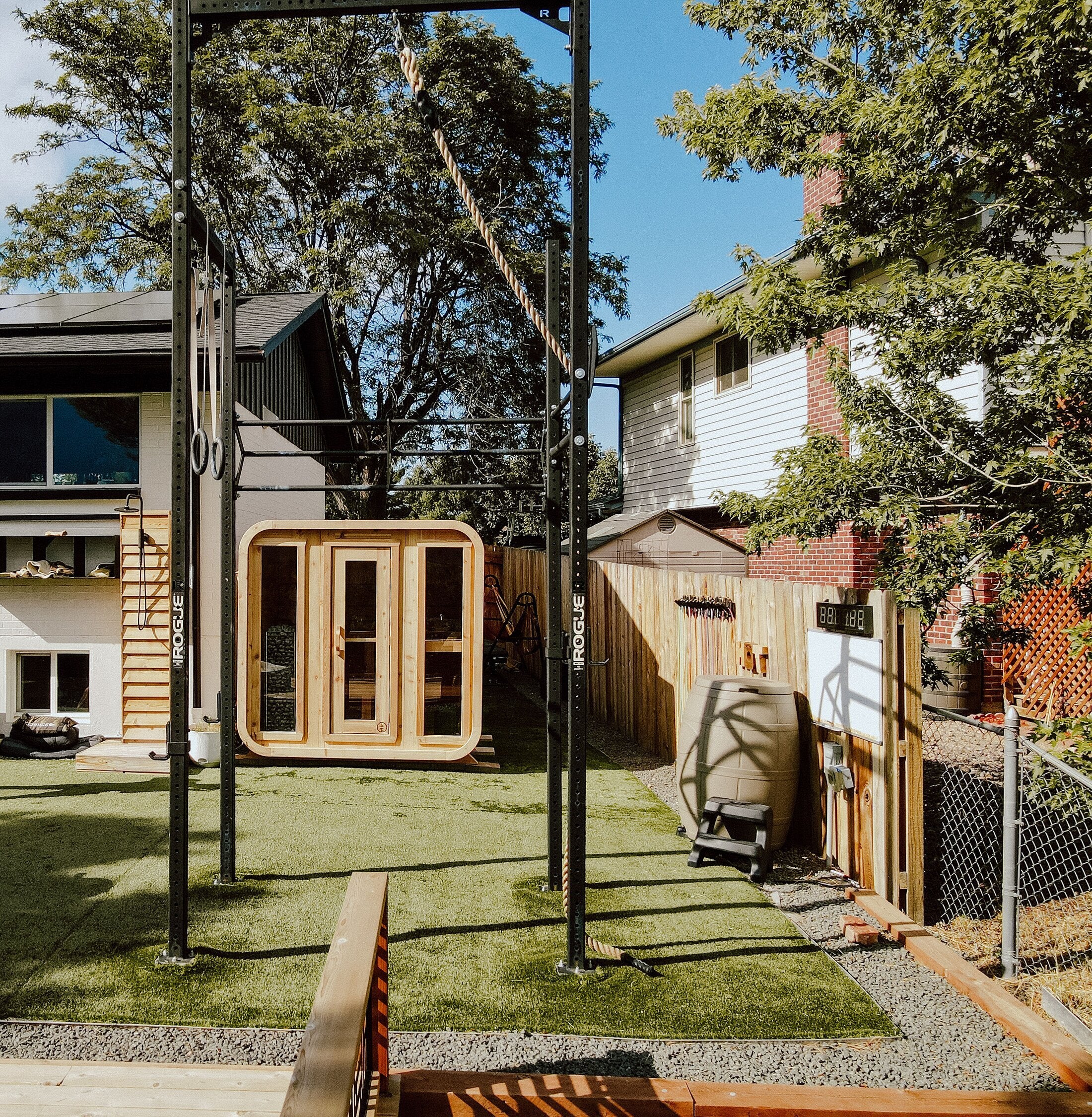 Cedar cabin sauna with three glass front panels in a turf backyard gym framed by a pull-up rig